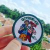 Ein Bild von einem Patch der von einer Hand mit schön lackierten Fingernägeln hochgehalten wird. Auf dem Ouddoor Kids Patch sind zwei Kinder aufgenäht.