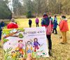 Gruppe von Erwachsenen steht im Wald auf einer Wiese im Kreis, im Vordergrund wird ein Buch mit dem Titel 'OUTDOOR-KIDS Praxishandbuch für Kindergruppen' gehalten.